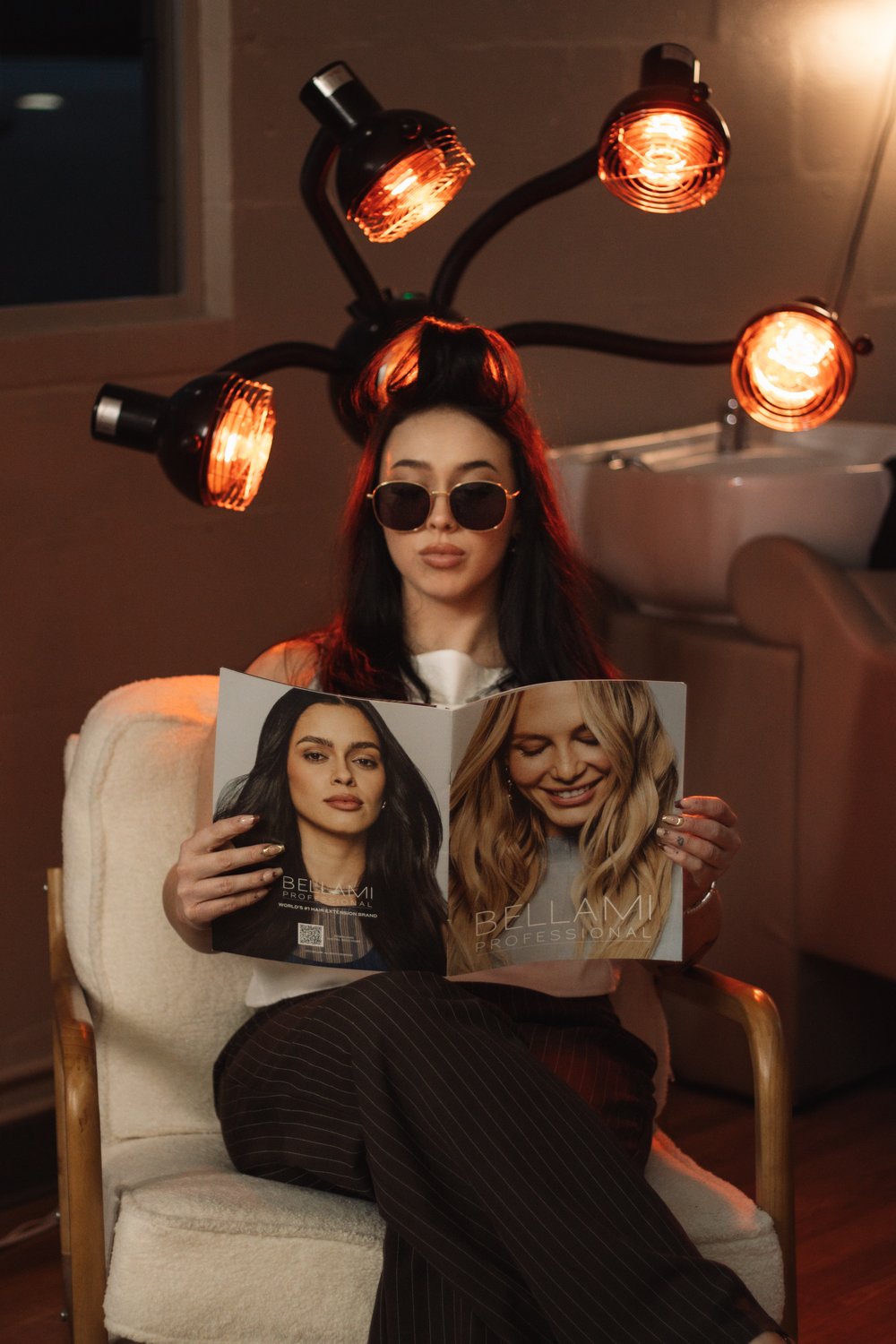 Stylist in sunglasses reading a hair magazine beneath glowing salon heat lamps