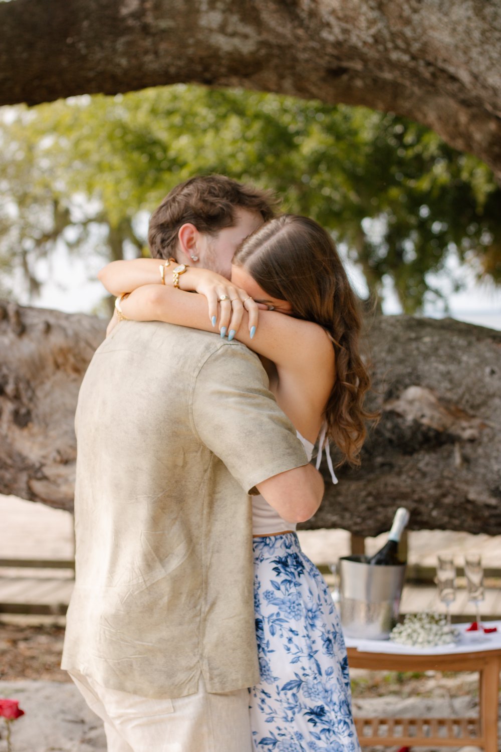Newly engaged couple hugging beside a champagne picnic under an oak