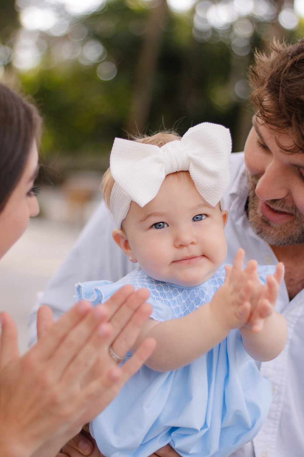 Baby girl in a big white bow clapping in her dad's arms as mom claps along