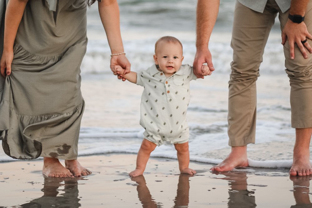 Toddler walking at the water's edge holding his parents' hands