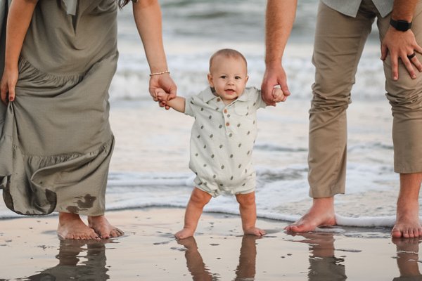Toddler walking at the water's edge holding his parents' hands
