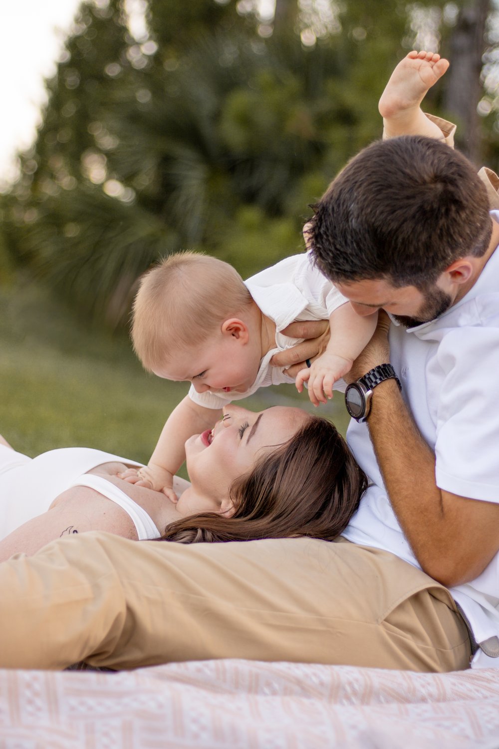 Dad lifting their laughing baby overhead while mom lies beside them on a blanket