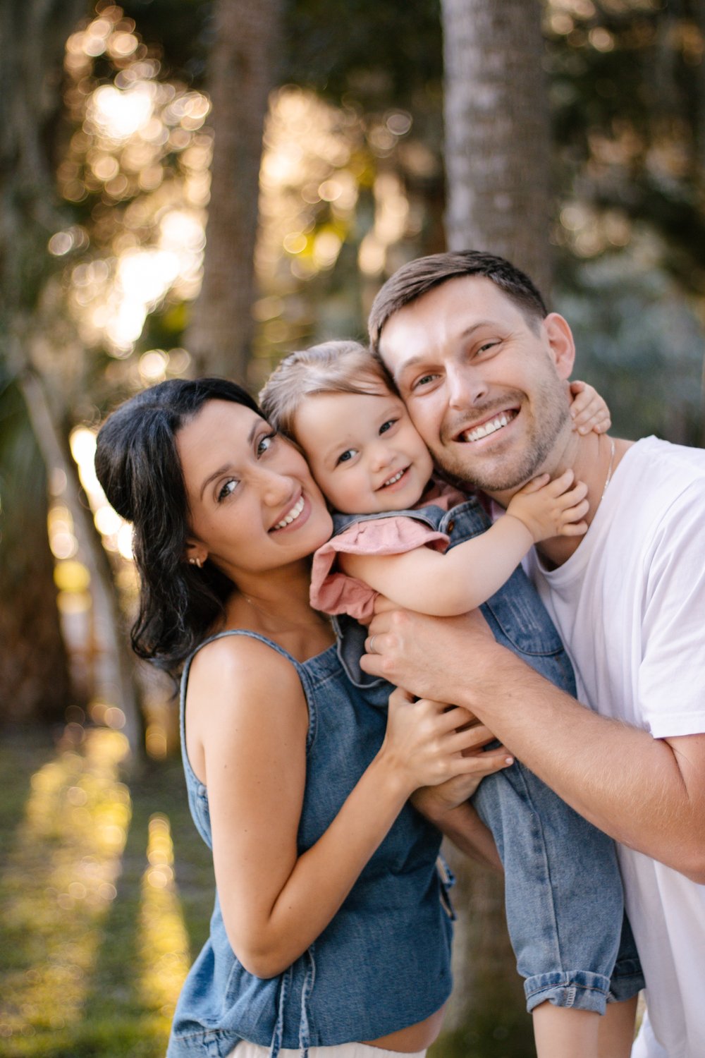 Mom and dad hugging their smiling toddler in denim overalls between them among the trees