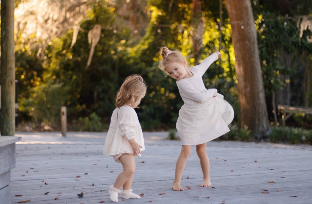 Two little girls in white dresses dancing on a wooden deck in warm evening light