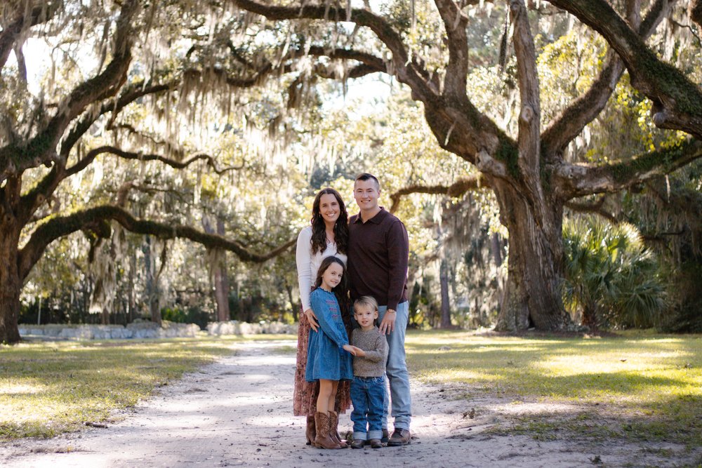 Family of four standing together on a sandy path under live oaks draped in Spanish moss