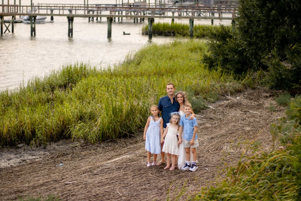 Family of five standing together on a marsh path by the water, with docks in the distance