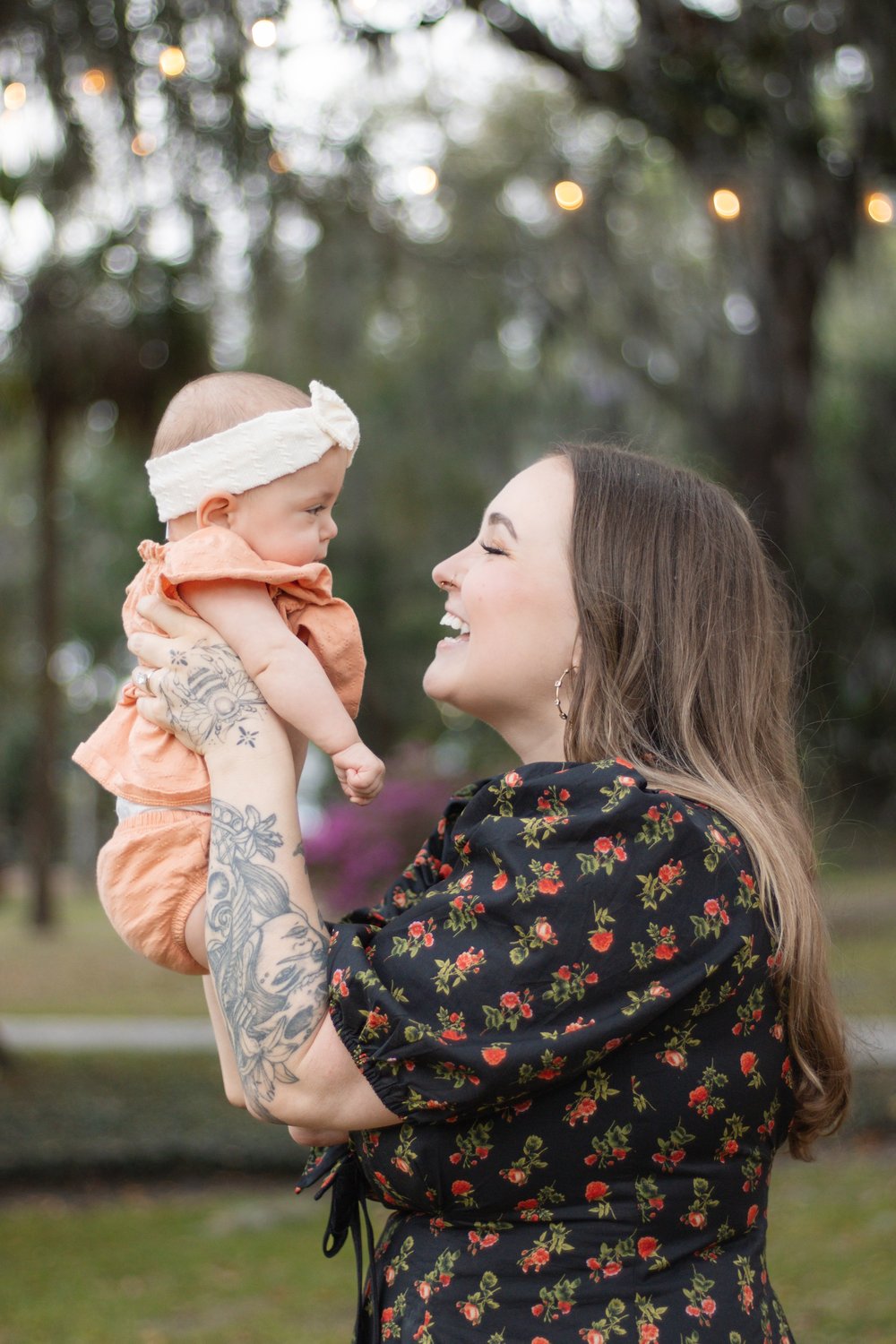 Laughing mother lifting her baby girl in a bow headband under twinkling string lights