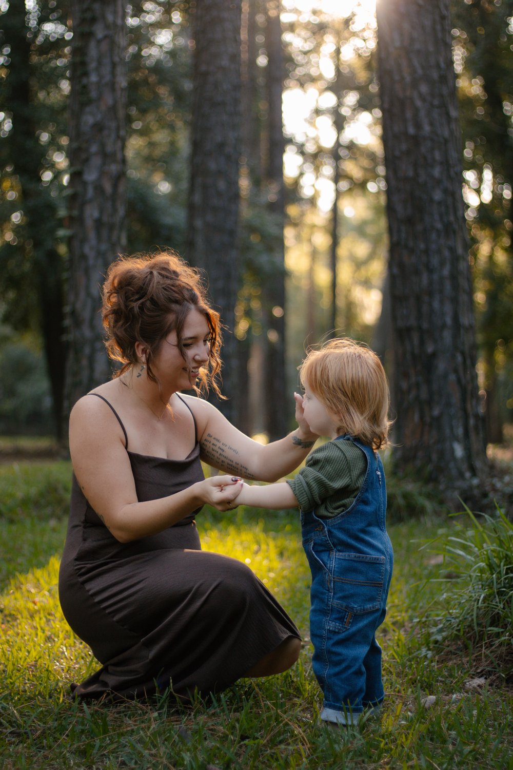 Mother kneeling to hold hands with her toddler in denim overalls in the pine woods