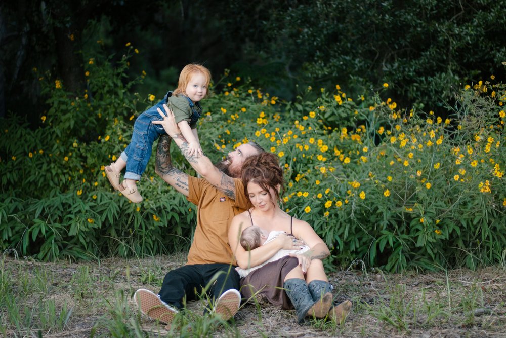 Dad lifting their toddler in overalls while mom holds their newborn in yellow wildflowers