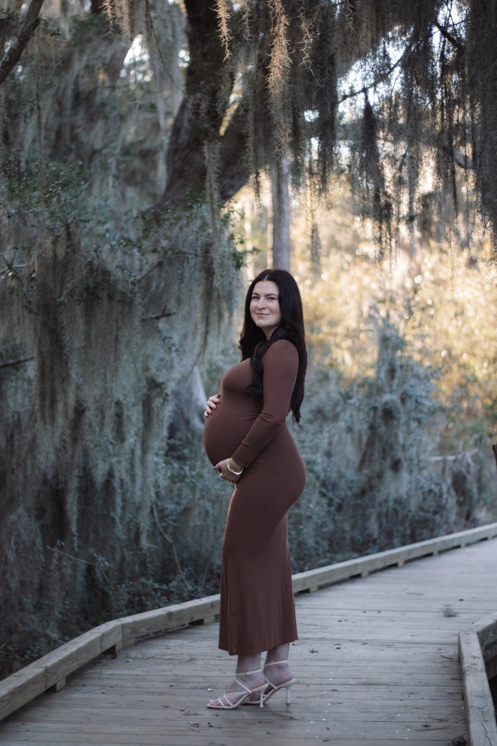 Expecting mother in a fitted brown dress on a wooden boardwalk beneath Spanish moss