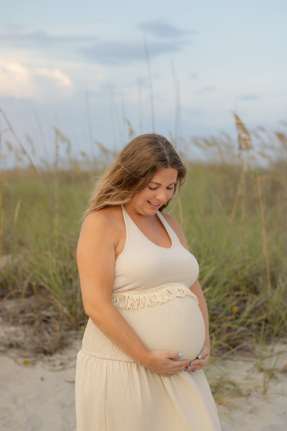 Expecting mother in a cream dress smiling down at her bump among the beach dunes and sea oats
