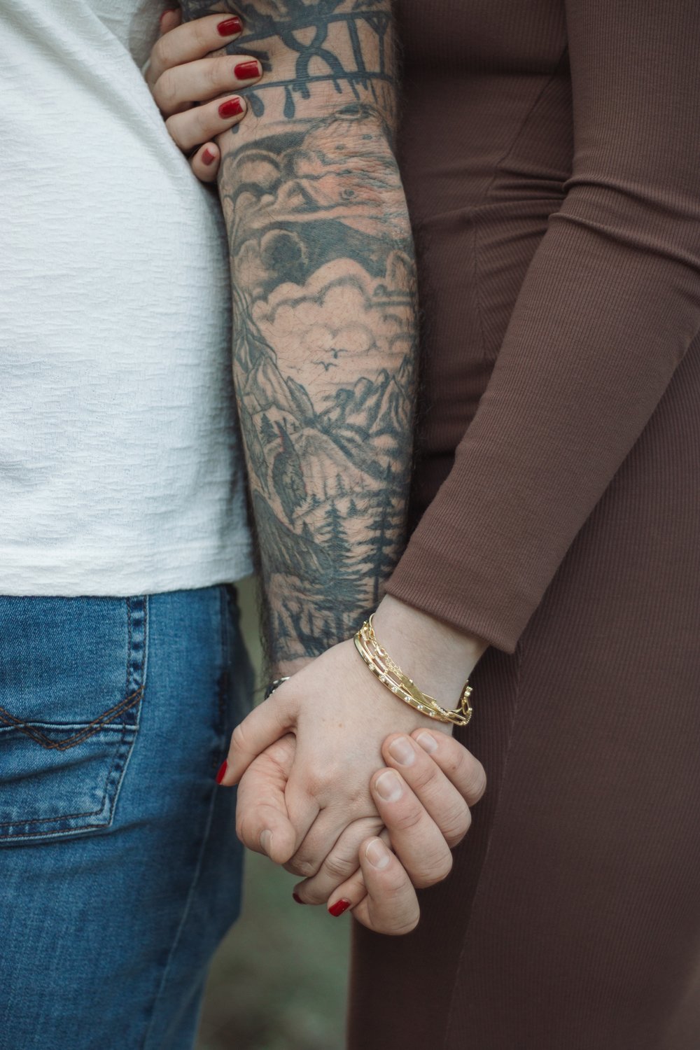 Close-up of an expecting couple holding hands, his tattooed arm beside her brown dress