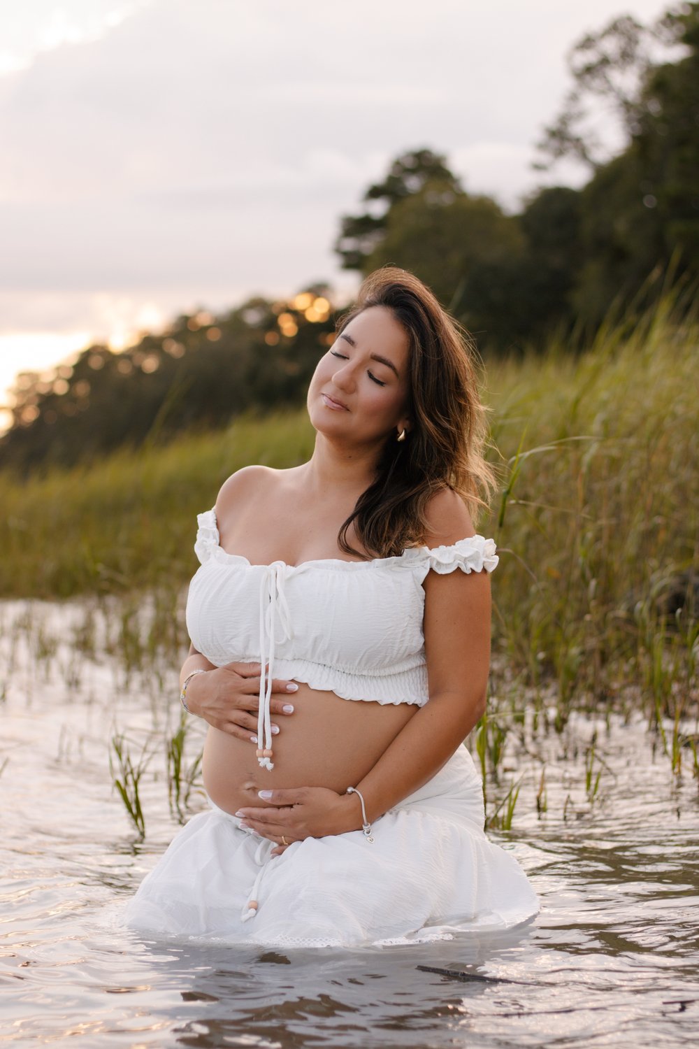 Expecting mother in a white dress kneeling in marsh water at sunset, hands on her bump