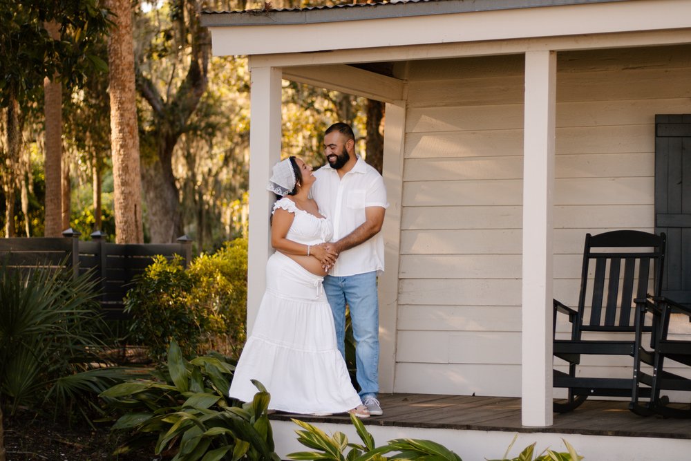 Expecting couple embracing on the porch of a white cottage