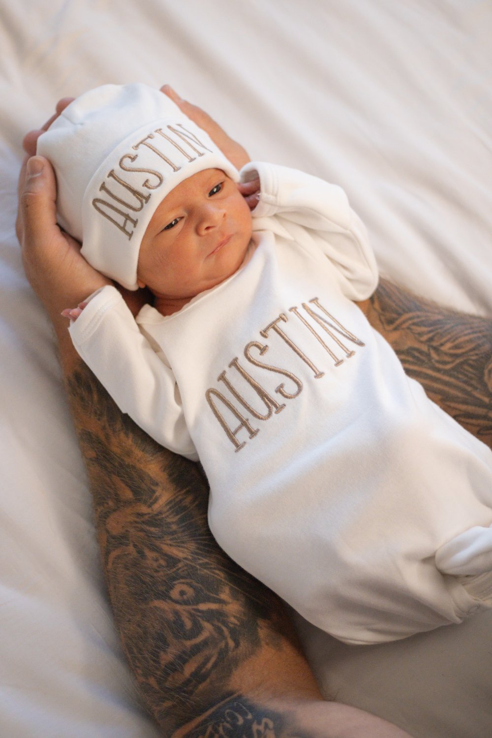 Newborn in a white hat and gown cradled in a parent's tattooed arms