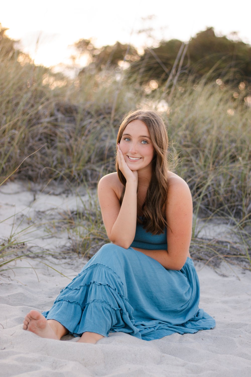 Graduate in a flowing blue dress sitting in the sand among the dunes