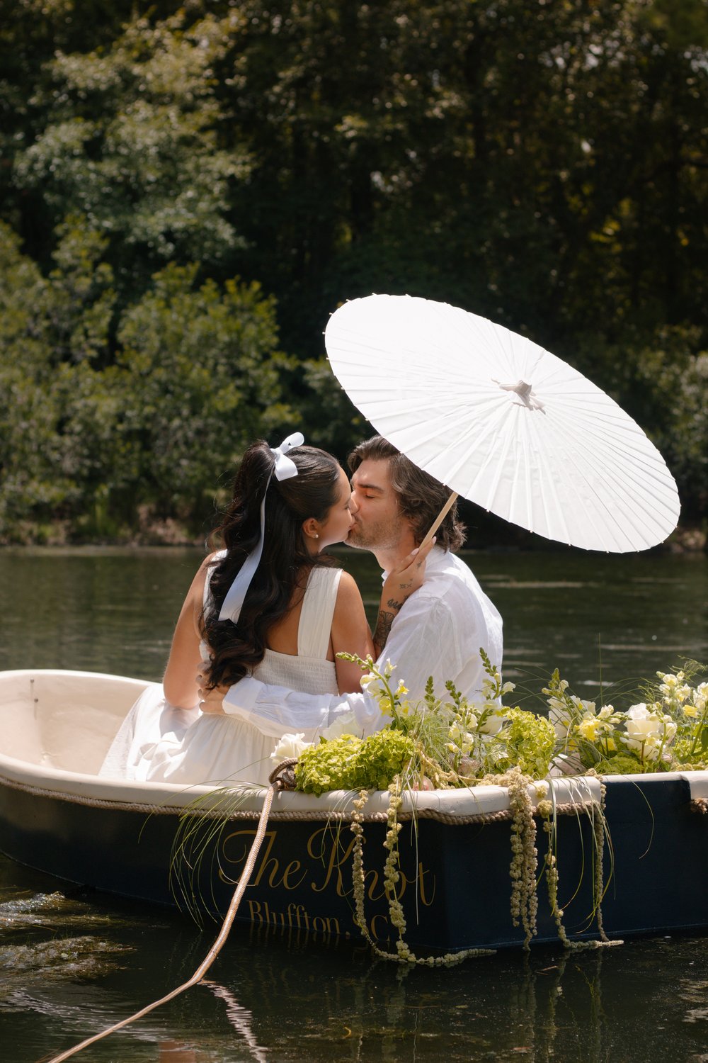 Couple sharing a kiss under a white parasol in a small flower-filled boat on the water